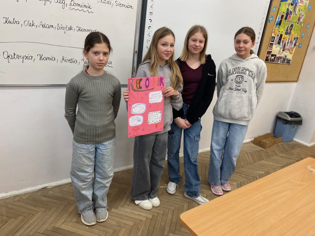 Klasa szkolna, tablica, ławki, tablica korkowa z plakatami, kosz na śmieci, czwórka dzieci, dziewczynki 12 lat ubrane w szare i czarne bluzy, dżinsy. Trzymają plakat.