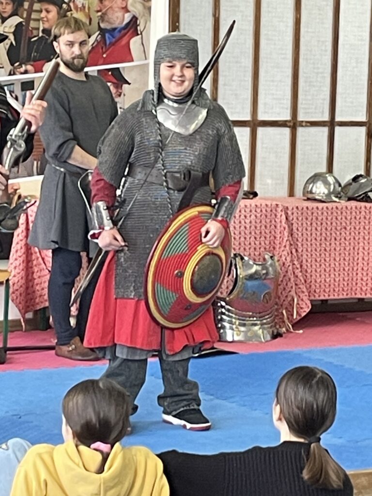 Na przedzie stoi chłopiec przebrany za żołnierza piechoty, ma na sobie kolczugę i inne elementy uzbrojenia.