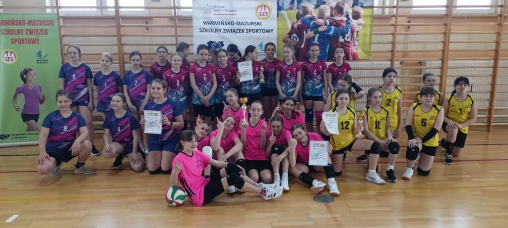Zdjęcie przedstawia grupę młodych dziewcząt, prawdopodobnie drużynę siatkówki, po turnieju lub zawodach. Dziewczynki są ułożone w kilku rzędach, trzymają dyplomy i puchary, co sugeruje sukces w zawodach. Na zdjęciu widoczne są numery na koszulkach zawodniczek, a także baner z logo Warmińsko-Mazurskiego Szkolnego Związku Sportowego. Ujęcia są zrobione w hali sportowej. Zdjęcie nadaje się do wykorzystania w kontekście: Sportu młodzieżowego: Idealne do stron internetowych klubów sportowych, szkół, lub organizacji promujących sport wśród dzieci i młodzieży. Sukcesów sportowych: Może być użyte na stronach prezentujących osiągnięcia sportowe, relacje z zawodów, lub galerii zdjęć. Promocji Warmińsko-Mazurskiego Szkolnego Związku Sportowego: Zdjęcie może być wykorzystane na stronie internetowej lub materiałach promocyjnych związku.