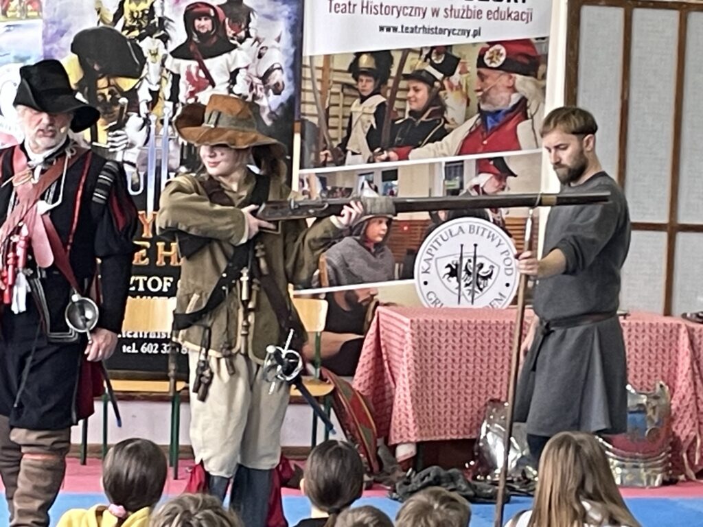 Na środku stoi dziewczyna przebrana za muszkietera, pokazuje strzał na podpórce.