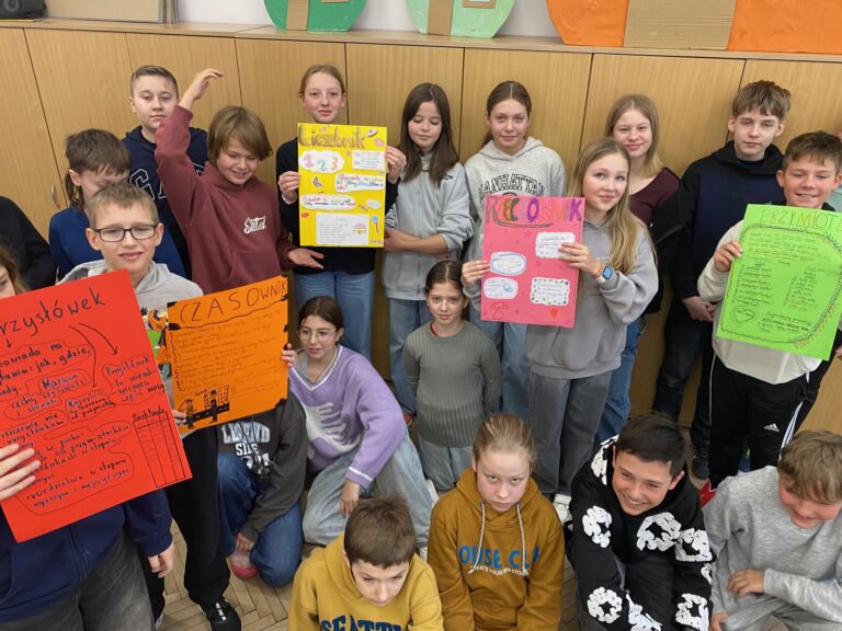 Klasa szkolna, szafy, osiemnaścioro dzieci, dziewczynki i chłopcy 12 lat ubrane w szare, czarne, żółte, granatowe, bordowe bluzy, trzymają kolorowe plakaty.