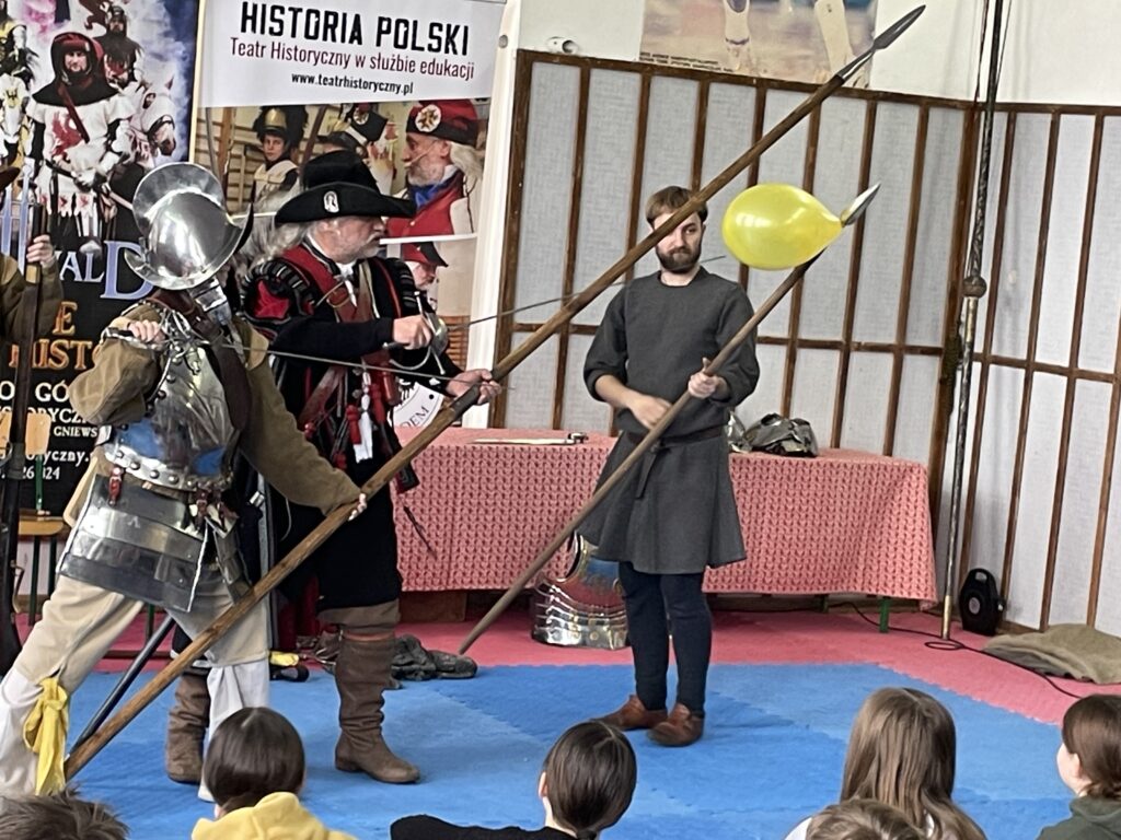 Na środku stoi chłopiec przebrany za pikiniera, pokazuje jak używać piki i rapiera. Uderza w balon.