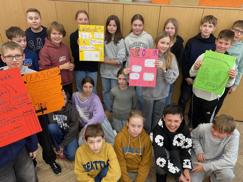 Klasa szkolna, szafy, dziewiętnaścioro dzieci, dziewczynki i chłopcy 12 lat ubrane w szare, czarne, żółte, granatowe, bordowe bluzy, trzymają kolorowe plakaty.