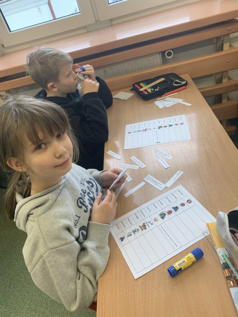 Przy ławce siedzi dwoje dzieci. Chłopiec patrzący przed siebie oraz dziewczynka, która uśmiech się do obiektywu. Oboje na ławce, przed sobą mają karty pracy i rozsypanki zdaniowe dotyczące wyrażeń mitologicznych, ich objaśnień i graficznego przedstawienia w formie obrazków.