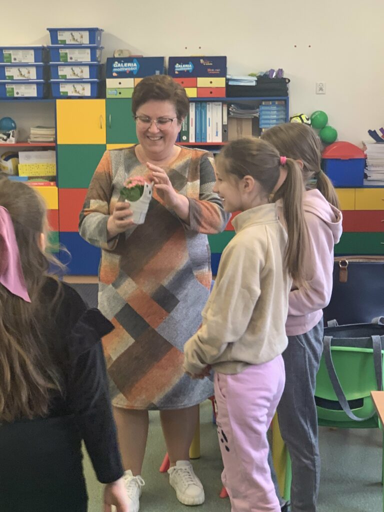 Na pierwszym planie widać panią Annę trzymającą w rękach kwiatek w doniczce w kształcie kotka. Pani Anna uśmiechnięta stoi w otoczeniu trójki dziewczynek, które przed chwilą podziękowały jej za przeprowadzone zajęcia i wręczyły ten prezent.