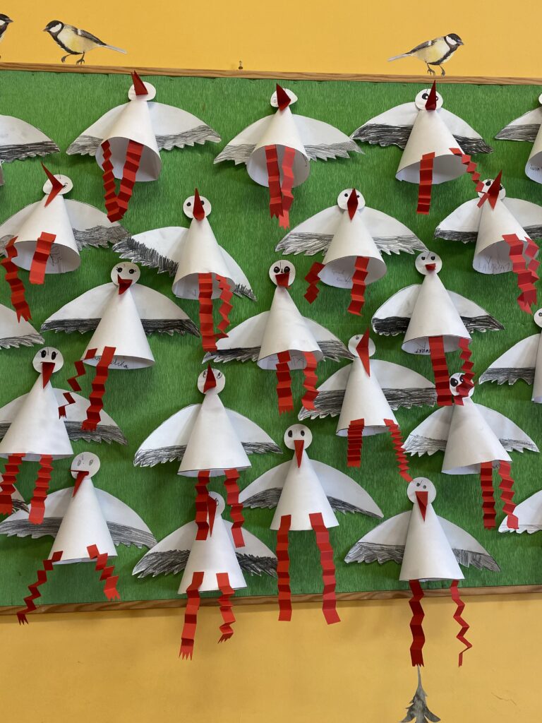 Na zdjęciu widać szkolną lub przedszkolną tablicę ozdobioną pracami plastycznymi przedstawiającymi bociany. Bociany zostały wykonane z papieru – ich tułowia mają kształt stożków, skrzydła są wycięte i pokolorowane na biało z szarymi elementami, a nogi i dzioby zrobione są z czerwonego papieru. Nogi zostały ozdobione w formie pasków zagiętych w harmonijkę, co nadaje im efekt trójwymiarowości. Tło tablicy jest zielone, a na górnej części umieszczono ilustracje sikorek, co dodaje kompozycji naturalnego wyglądu. Całość sprawia wrażenie starannie przygotowanej dekoracji, prawdopodobnie nawiązującej do tematu wiosny i powrotu bocianów.
