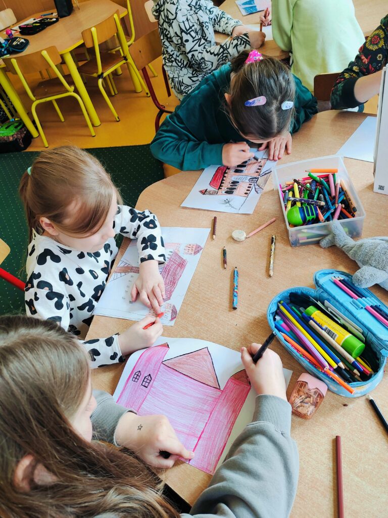 Na zdjęciu widać grupę dzieci siedzących przy stole i rysujących lub kolorujących obrazki przedstawiające budynki. Na stole znajdują się przybory plastyczne, takie jak kredki, ołówki, temperówki i gumki do mazania. Dzieci są skupione na swojej pracy, używając różnych kolorów, a niektóre mają ozdobne spinki we włosach. W tle widać inne dzieci przy sąsiednim stole oraz żółte krzesła i stoliki typowe dla szkolnej klasy.