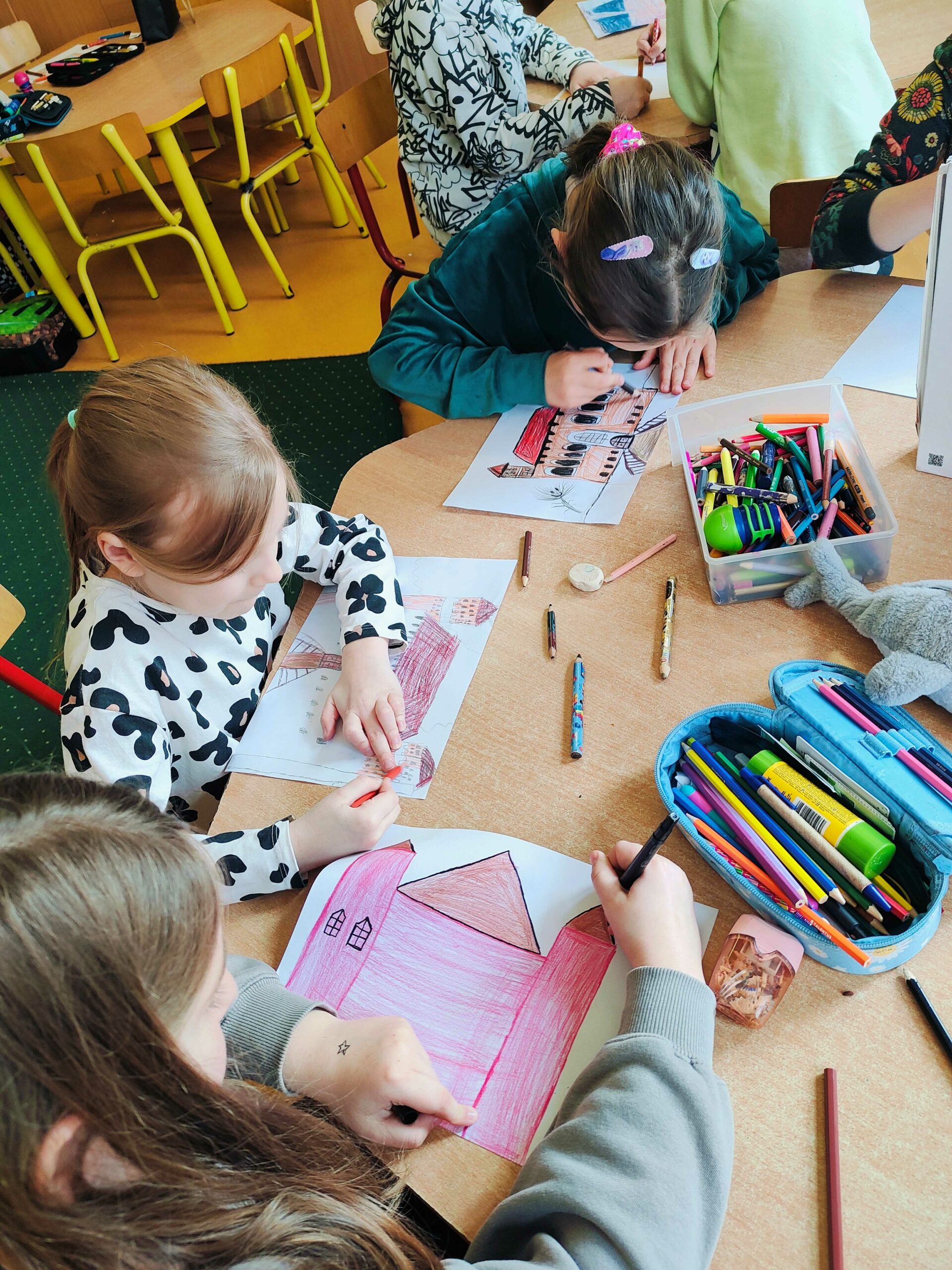 Na zdjęciu widać grupę dzieci siedzących przy stole i rysujących lub kolorujących obrazki przedstawiające budynki. Na stole znajdują się przybory plastyczne, takie jak kredki, ołówki, temperówki i gumki do mazania. Dzieci są skupione na swojej pracy, używając różnych kolorów, a niektóre mają ozdobne spinki we włosach. W tle widać inne dzieci przy sąsiednim stole oraz żółte krzesła i stoliki typowe dla szkolnej klasy.