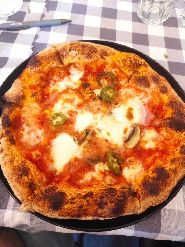 Zdjęcie przedstawia pizzę na czarnym talerzu, znajdującą się na stole z biało-szarą kratkowaną serwetą. Pizza ma chrupiące, lekko przypieczone brzegi i jest pokryta sosem pomidorowym, roztopionym serem mozzarella oraz dodatkami, takimi jak pieczarki i plasterki zielonej papryczki. W tle widać szklankę, długopis i inne elementy stołu.