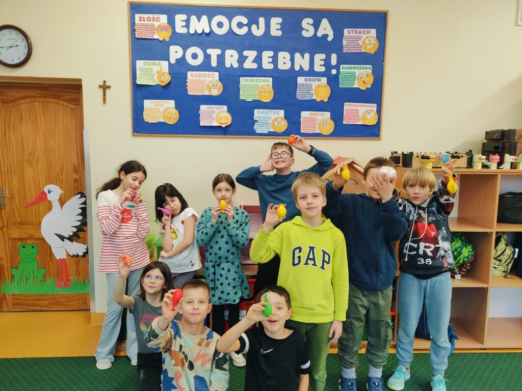 Na zdjęciu znajduje się grupa dzieci w klasie szkolnej, prezentujących kolorowe, ręcznie wykonane przedmioty, prawdopodobnie sensoryczne gniotki. W tle widać tablicę z napisem "EMOCJE SĄ POTRZEBNE!" oraz ilustracjami przedstawiającymi różne emocje, co sugeruje, że dzieci mogły brać udział w zajęciach dotyczących emocji. Pomieszczenie jest przyjaźnie urządzone, z dekoracjami na drzwiach, półkami na plecaki oraz zielonym dywanem na podłodze. Dzieci wyglądają na zadowolone i dumne ze swoich prac, pozując radośnie do zdjęcia.