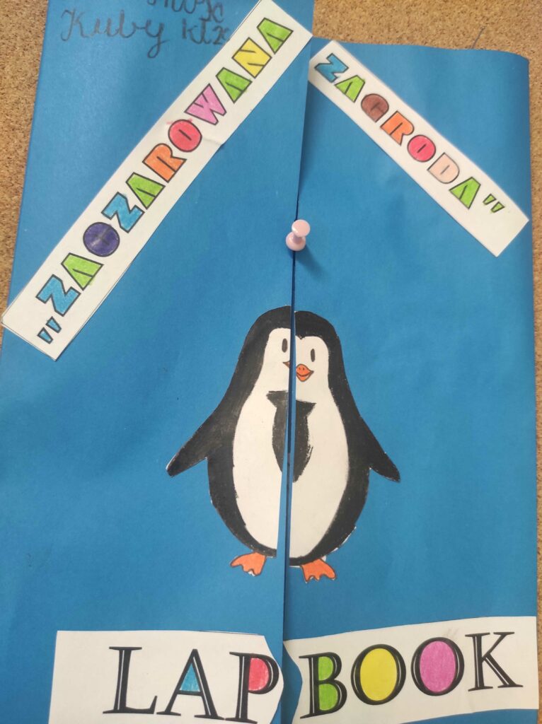 Na zdjęciu widać niebieską teczkę lub okładkę lapbooka (książki interaktywnej) zatytułowanego "ZAOCZAROWANA ZAGRODA". Okładka jest ozdobiona rysunkiem pingwina, który został podzielony na dwie części, ponieważ okładka otwiera się na środku. Tytuł jest napisany kolorowymi literami na białych paskach papieru, a na dole widnieje napis "LAPBOOK", również w kolorowej czcionce.Lapbook dotyczy książki "Zaczarowana zagroda" autorstwa Aliny i Czesława Centkiewiczów, która opowiada o pingwinach i ich życiu na Antarktydzie.