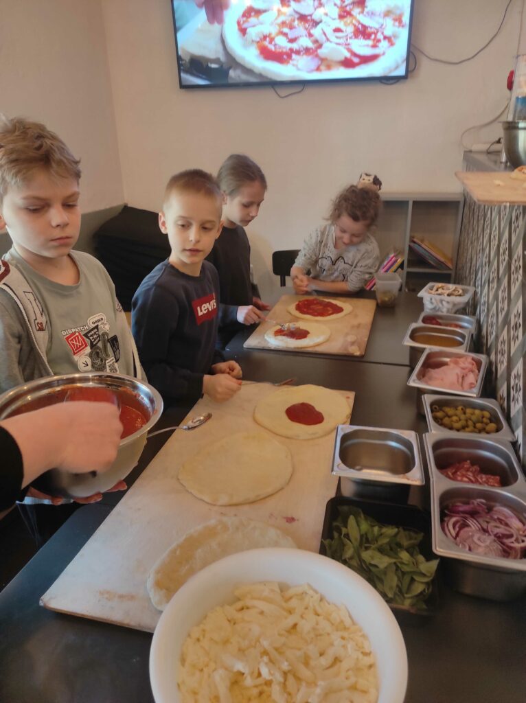 Zdjęcie pokazuje grupę dzieci biorących udział w warsztatach kulinarnych, gdzie przygotowują własne pizzę. Są skupione na rozprowadzaniu sosu pomidorowego na surowym cieście. Wokół nich znajduje się stół z różnymi składnikami, takimi jak ser, szynka, oliwki, cebula, bazylia i inne dodatki. W tle na ścianie wisi ekran wyświetlający obraz pizzy.