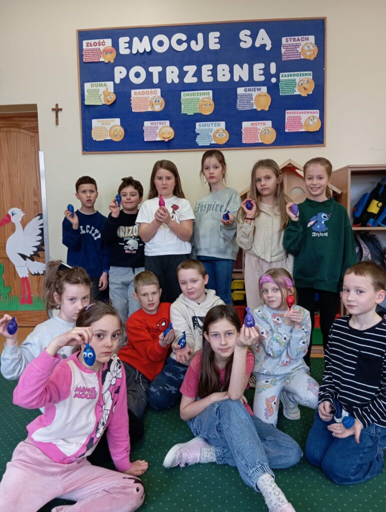 Na zdjęciu znajduje się grupa dzieci w klasie, które prezentują własnoręcznie wykonane sensoryczne gniotki w różnych kolorach. W tle widoczna jest tablica edukacyjna z napisem "EMOCJE SĄ POTRZEBNE!" oraz opisami różnych emocji, co sugeruje, że zajęcia dotyczyły uczuć i ich wyrażania. Dzieci mają poważne, ale także zaciekawione miny, a ich postawa wskazuje na zaangażowanie w temat. Sala jest kolorowa i przyjazna, co tworzy ciepłą atmosferę sprzyjającą nauce i zabawie.
