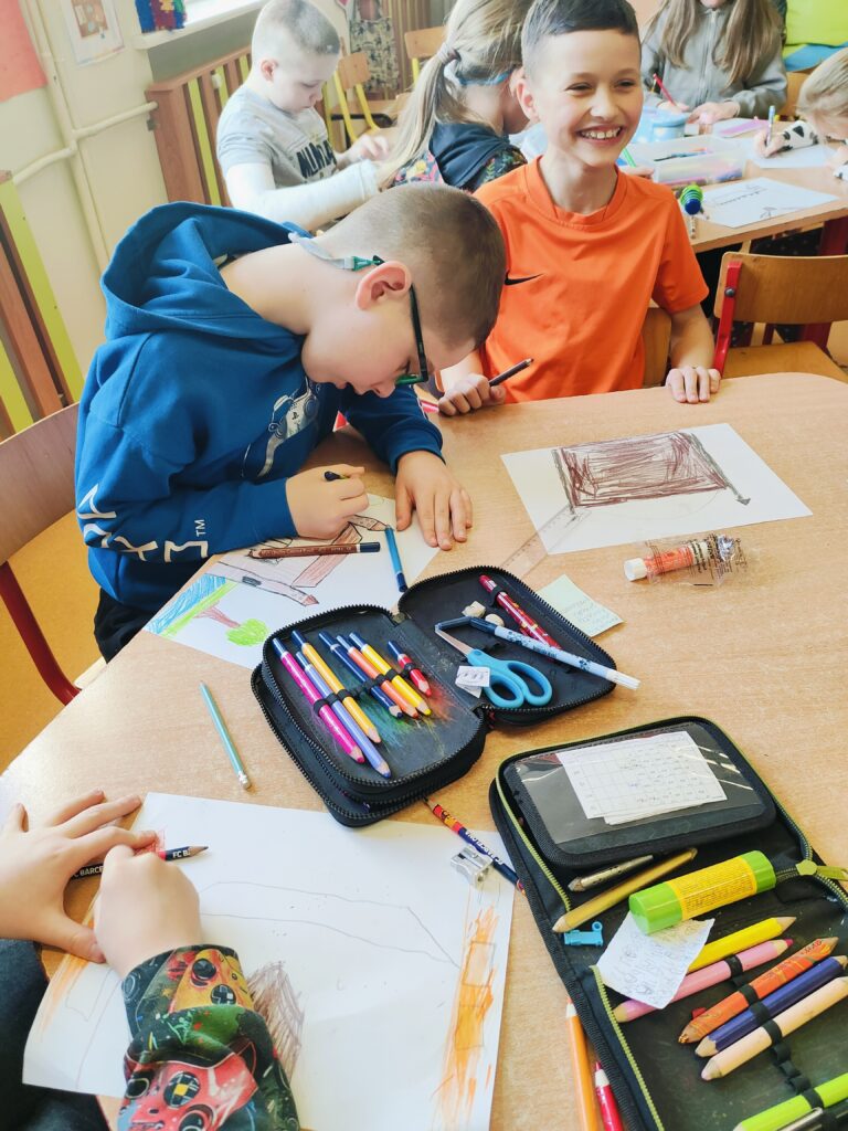 Na zdjęciu widać dzieci pracujące nad rysunkami w klasie szkolnej. Chłopiec w niebieskiej bluzie pochyla się nad swoim rysunkiem, podczas gdy chłopiec w pomarańczowej koszulce uśmiecha się szeroko. Na stole leżą piórniki pełne kredek, ołówków, nożyczek i kleju, a dzieci kolorują swoje prace. W tle inne dzieci również rysują, a sala ma jasne ściany i drewniane meble, co tworzy ciepłą atmosferę.