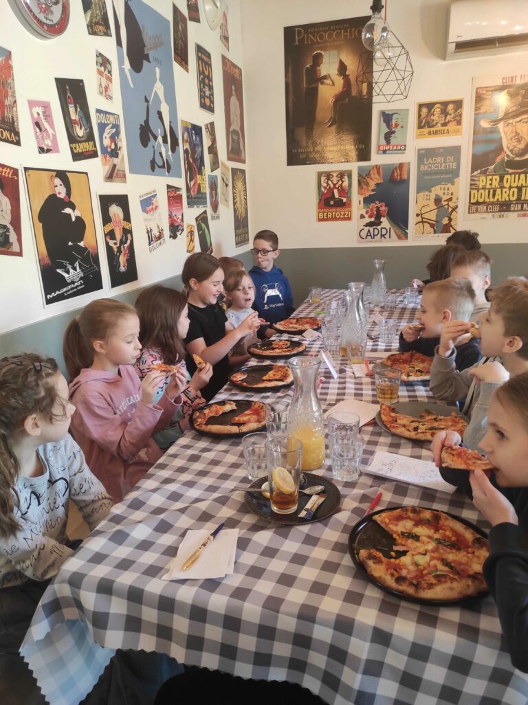 Zdjęcie przedstawia grupę dzieci siedzących przy długim stole w restauracji w stylu włoskim. Stół jest przykryty obrusami w biało-szarą kratę, a na nim znajdują się talerze z pizzą, szklanki z napojami, karafki z sokiem oraz przybory do rysowania. Dzieci, ubrane w codzienne stroje, jedzą pizzę i rozmawiają ze sobą, wyglądając na zadowolone. Ściany lokalu ozdobione są licznymi plakatami w stylu retro, nawiązującymi do włoskiego kina i kultury, m.in. widać plakaty filmowe, reklamy włoskich produktów i grafiki artystyczne.