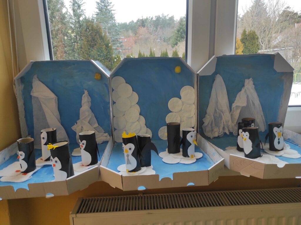 Na zdjęciu widoczne są makiety przedstawiające zimowy, antarktyczny krajobraz z pingwinami. Zostały one wykonane z kartonowych pudełek po pizzy, które służą jako tło i podstawa dla scenek.Opis elementów makiety:Tło zostało pomalowane na niebiesko, co symbolizuje niebo. Dodatkowo znajdują się na nim białe elementy wykonane z bibuły lub waty, przedstawiające lodowce i góry lodowe. Pingwiny zostały wykonane z rolek po papierze toaletowym owiniętych czarnym papierem. Mają naklejone białe brzuszki, narysowane oczy oraz wycięte z papieru dzióbki i skrzydła. Niektóre pingwiny mają dodatkowe ozdoby, np. żółtą koronę lub krawat.Lód i śnieg zostały stworzone z kawałków białego papieru i wacików kosmetycznych, które nadają teksturę śnieżnym krajobrazom.Słońce symbolizuje żółta kuleczka w rogu jednej z makiet, co dodaje pracy trochę ciepła.Makiety są ustawione na parapecie okiennym, a całość wygląda jak kreatywny projekt edukacyjny o Antarktydzie i pingwinach.