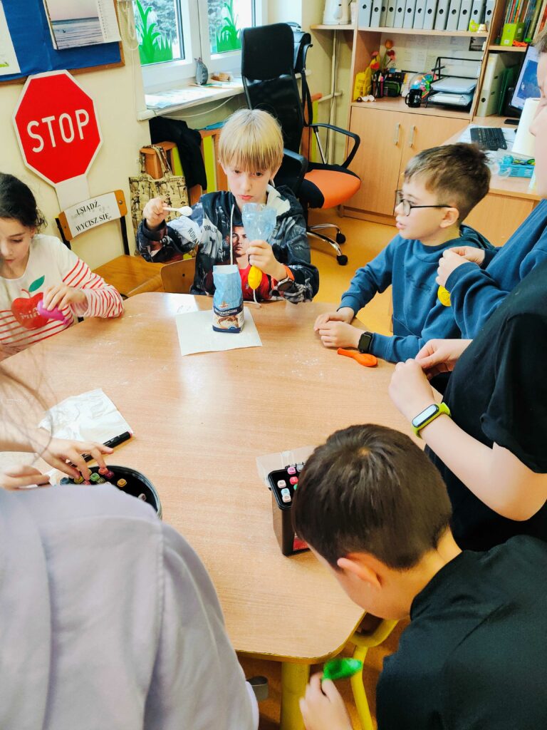 Na zdjęciu widać grupę dzieci przy stole, które biorą udział w kreatywnych zajęciach. W centrum uwagi jest chłopiec napełniający balon mąką przy użyciu lejka, co sugeruje, że dzieci tworzą sensoryczne gniotki. Wokół stołu znajdują się różne materiały, takie jak pisaki i inne przybory, a w tle widać klasę z biurkiem nauczyciela i oznakowaniem "STOP" oraz "USIĄDŹ, WYCISZ SIĘ". Dzieci są zaangażowane w swoją pracę, a atmosfera wydaje się pełna skupienia i twórczej zabawy.