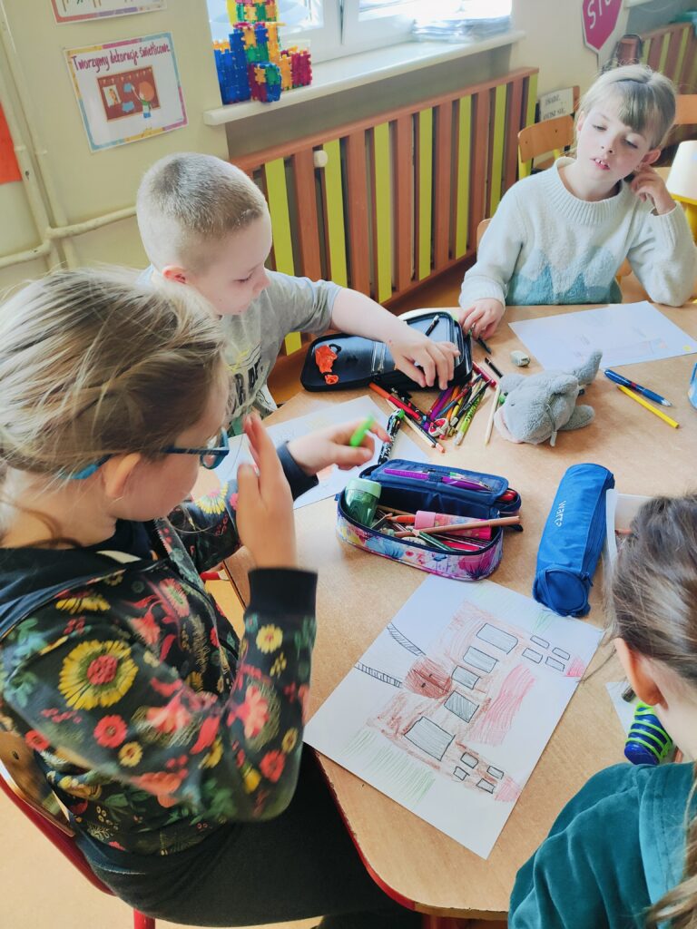 Na zdjęciu widać grupę dzieci siedzących przy stole i zajmujących się rysowaniem oraz kolorowaniem obrazków przedstawiających budynki. Na stole leżą różne przybory szkolne, takie jak kredki, piórniki, gumki do mazania oraz pluszowa zabawka. Dzieci są skupione na pracy, a jedno z nich, w kolorowej bluzie, poprawia okulary lub włosy. W tle widać klasę szkolną z kolorowymi plakatami, klockami oraz oknem, przez które wpada naturalne światło.