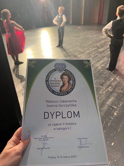 Na zdjęciu widać dyplom trzymany w dłoni przez osobę. Dyplom został przyznany Mateuszowi Sałamacha i Joannie Gorczyńskiej za zajęcie V miejsca w kategorii I podczas Ogólnopolskiego Konkursu o „Pierścień Księżnej Izabeli”. W tle na scenie występują dzieci ubrane w stroje taneczne – dziewczynka w czerwonej sukience, a chłopcy w białych koszulach i kamizelkach.