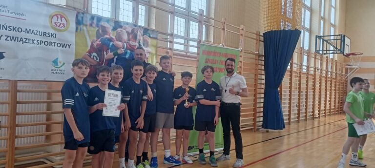 Zdjęcie przedstawia grupę młodych chłopców, prawdopodobnie drużynę sportową, stojących w hali sportowej po zawodach. Chłopcy trzymają dyplomy i puchar, co sugeruje, że zajęli wysokie miejsce w turnieju. Uśmiechnięci i dumni, stoją obok trenera, który również wygląda na zadowolonego. W tle widoczne są banery z logo organizacji sportowej, co wskazuje na oficjalny charakter wydarzenia. Atmosfera na zdjęciu jest radosna i pełna pozytywnych emocji, związanych z sukcesem sportowym. Chłopcy są ubrani w jednakowe koszulki klubowe, co podkreśla ich przynależność do drużyny. Zdjęcie jest dobrze naświetlone i ostre, co pozwala na wyraźne dostrzeżenie szczegółów.