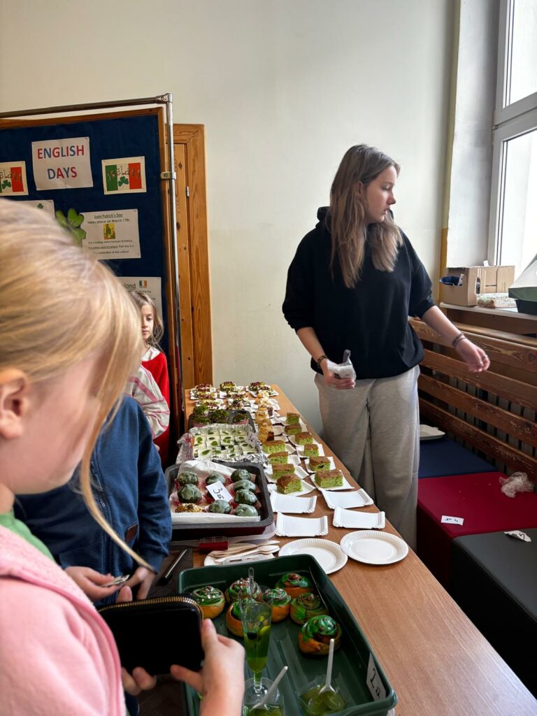 Na zdjęciu widzimy stoisko z jedzeniem. Na stole znajdują się różne zielone wypieki i słodycze, a kilka osób, w tym dzieci, ogląda i kupuje smakołyki. W tle widać tablicę z napisem “ENGLISH DAYS” oraz informacje o Irlandii i Dniu Świętego Patryka. Przy stoisku stoi młoda dziewczyna w czarnej bluzie i jasnych spodniach, pomagająca w sprzedaży.