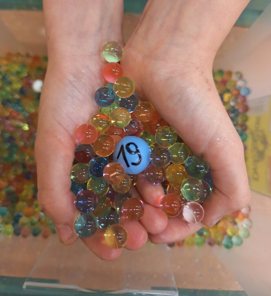 Dziecko trzyma w dłoniach kolorowe kulki żelowe, wśród których znajduje się jedna większa, niebieska, z liczbą „19”. Aktywność polega na wyszukiwaniu oznaczonych kulek, które wskazują zadania do wykonania, łącząc zabawę sensoryczną z nauką.