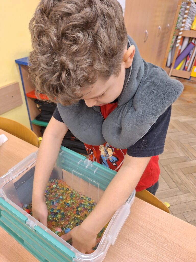 Chłopiec z lokami zanurza ręce w plastikowym pojemniku pełnym kolorowych hydrożelowych kulek. Na jego szyi spoczywa obciążający kołnierz sensoryczny. Znajduje się w klasie, gdzie w tle widoczne są szafki z pomocami dydaktycznymi.