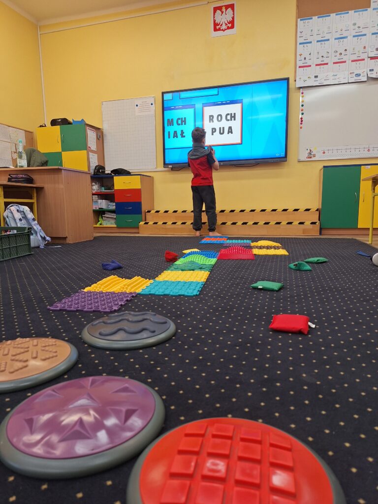 Zdjęcie przedstawia dziecko chodzące boso po sensorycznych krążkach dotykowych o różnych fakturach i kolorach. W tle znajduje się sala lekcyjna z dywanem, na którym ułożono dodatkowe ścieżki sensoryczne. Na ekranie widoczna jest tablica interaktywna z literami do odczytania.