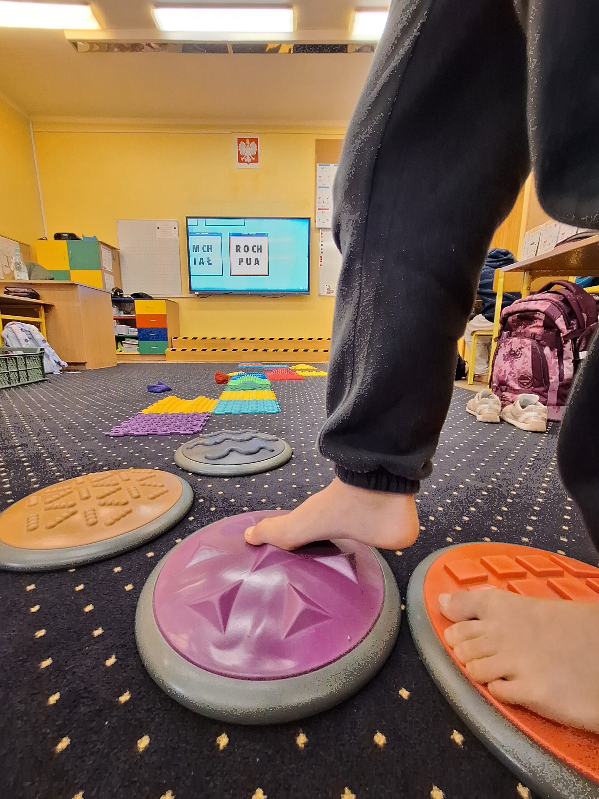 Uczeń przechodzi boso po ścieżce sensorycznej złożonej z kolorowych, fakturowanych dysków i mat, rozwijając zmysł dotyku i równowagę. W tle widać tablicę multimedialną z zadaniami ortograficznymi dotyczącymi wyrazów z „ch”, co wskazuje na połączenie aktywności sensorycznej z nauką języka polskiego.