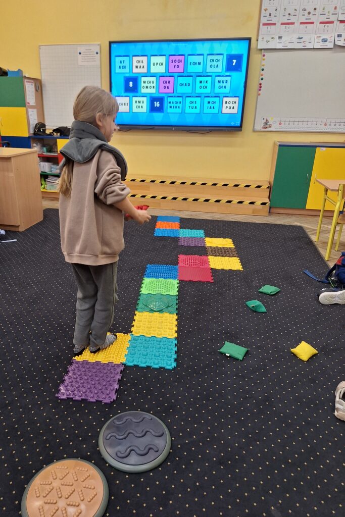 Dziewczynka w kołnierzu obciążeniowym przechodzi po kolorowej ścieżce sensorycznej, rozwijając koordynację i percepcję dotykową. W tle widoczna jest tablica multimedialna z zadaniami ortograficznymi dotyczącymi wyrazów z „ch”, co łączy aktywność ruchową z nauką języka polskiego.