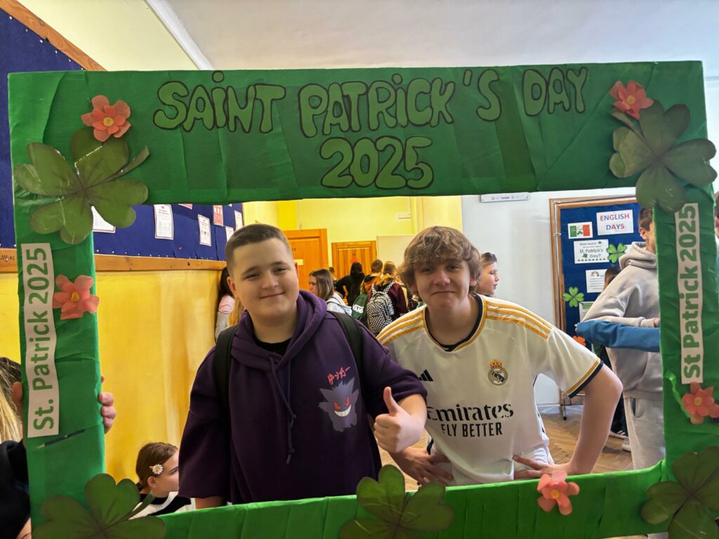 Na zdjęciu trzech chłoców pozuje do zdjęcia, trzymając dużą zieloną ramkę z napisem „SAINT PATRICK’S DAY 2025”. Ramka jest ozdobiona koniczynkami i kwiatami, a także ma boczne paski z napisem „St. Patrick 2025”. Chłopcy są uśmiechnięci, a w tle widać szkolny korytarz z innymi uczniami.