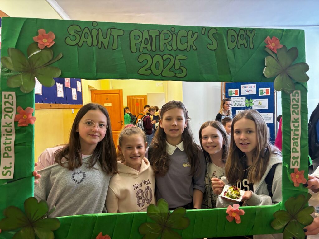Na zdjęciu 5 dziewczyn pozuje do zdjęcia, trzymając dużą zieloną ramkę z napisem „SAINT PATRICK’S DAY 2025”. Ramka jest ozdobiona koniczynkami i kwiatami, a także ma boczne paski z napisem „St. Patrick 2025”. Dziewczyny są uśmiechnięte, a w tle widać szkolny korytarz z innymi uczniami.