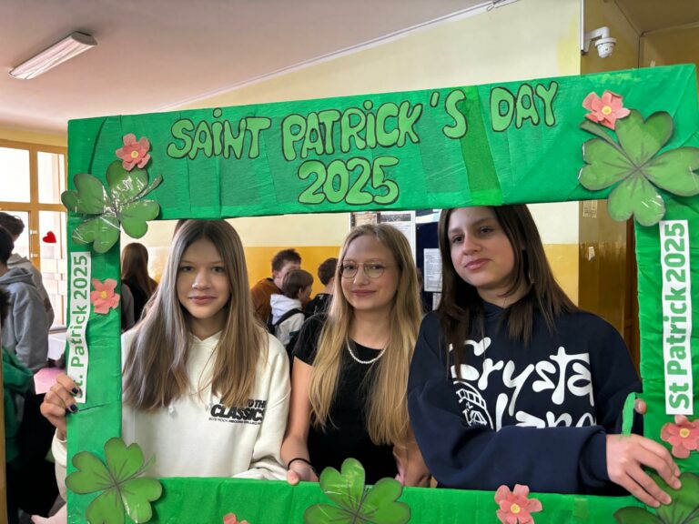 Na zdjęciu trzy dziewczyny pozują do zdjęcia, trzymając dużą zieloną ramkę z napisem „SAINT PATRICK’S DAY 2025”. Ramka jest ozdobiona koniczynkami i kwiatami, a także ma boczne paski z napisem „St. Patrick 2025”. Dziewczyny są uśmiechnięte, a w tle widać szkolny korytarz z innymi uczniami.