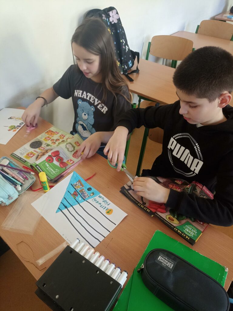 W centrum uwagi uczniów klasy 4c znajduje się gazetka, na której widać grupy produktów spożywczych, takie jak owoce, warzywa, zboża. Chłopiec wycina ilustracje przedstawiające grupy produktów spożywczych.