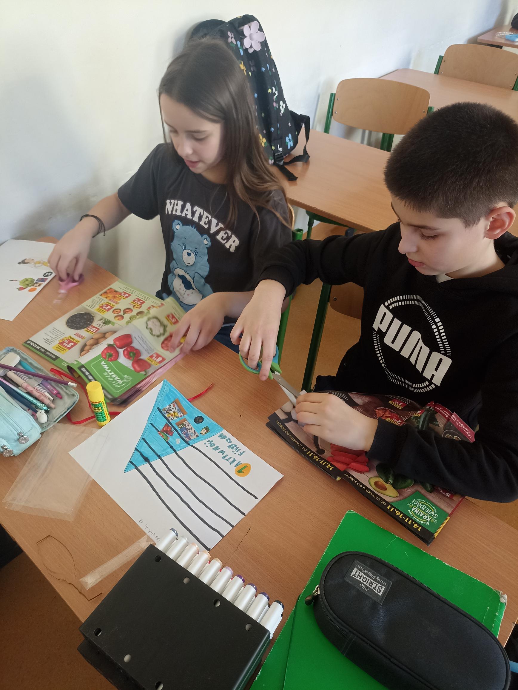 W centrum uwagi uczniów klasy 4c znajduje się gazetka, na której widać grupy produktów spożywczych, takie jak owoce, warzywa, zboża. Chłopiec wycina ilustracje przedstawiające grupy produktów spożywczych.