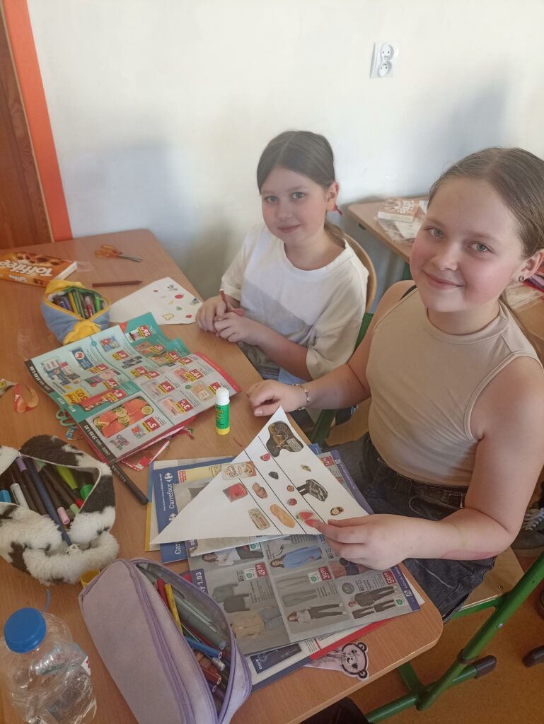 Na stole obok uczennic klasy 4c leżą kolorowe flamastry, nożyczki i klej, co wskazuje na to, że dzieci są w pełni zaangażowane w proces twórczy. Atmosfera jest radosna i kreatywna, a dzieci uczą się, jak ważne jest zdrowe żywienie w zabawny i interaktywny sposób.