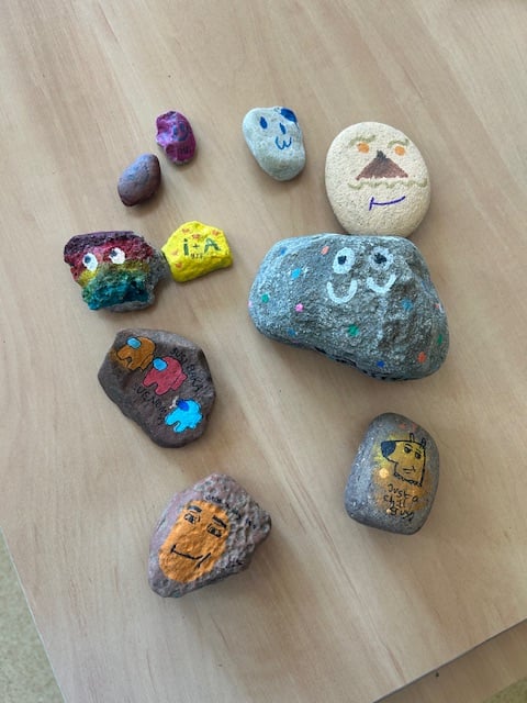 Na zdjęciu widzimy kolekcję pomalowanych kamyków ułożonych na drewnianym stole. Każdy z nich ma unikalne wzory i kolory, a niektóre przedstawiają uśmiechnięte buźki, postacie oraz kreatywne abstrakcyjne wzory. Kamienie są różnej wielkości i kształtu – niektóre są gładkie i jasne, inne bardziej chropowate i ciemniejsze. Widać, że zostały pomalowane z dużą starannością i kreatywnością, prawdopodobnie przez dzieci w ramach twórczego projektu. Całość sprawia wrażenie radosnej i pozytywnej inicjatywy, mającej na celu wywołanie uśmiechu u osób, które znajdą te wyjątkowe kamienie.