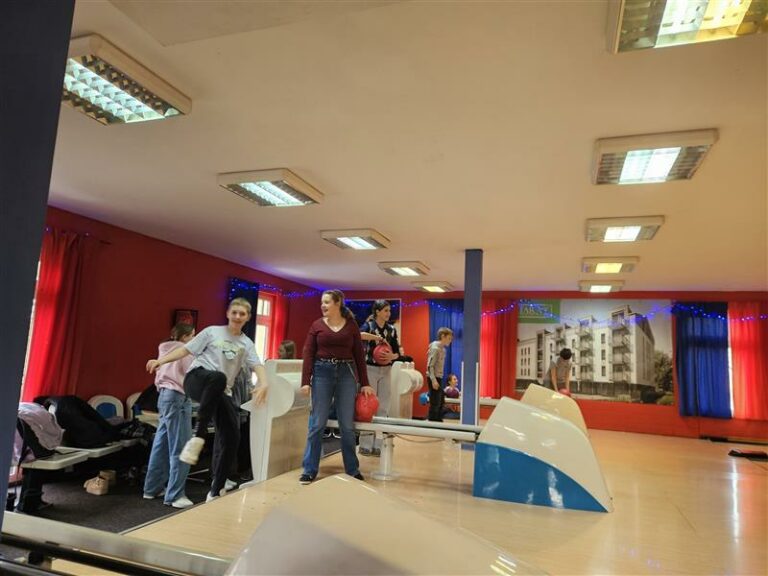 Na zdjęciu widać grupę młodzieży bawiącą się na torze do kręgli. Pomieszczenie jest przytulne i kolorowe – czerwone ściany, niebieskie zasłony i dekoracyjne światełka tworzą wesołą atmosferę. W tle znajduje się duży plakat z wizerunkiem nowoczesnego budynku, co dodaje ciekawego akcentu wystrojowi. Na pierwszym planie widzimy dziewczynkę i chłopca – dziewczyna w bordowej bluzce i jeansach trzyma czerwoną kulę, gotowa do rzutu, chłopiec w szarej koszulce i czarnych spodniach radośnie podskakuje, wyraźnie ciesząc się grą. W tle inni uczestnicy rozgrywki również są zaangażowani – jedni przygotowują się do rzutu, inni kibicują lub rozmawiają. Zdjęcie oddaje atmosferę świetnej zabawy, integracji i sportowej rywalizacji, co czyni ten dzień wyjątkowym