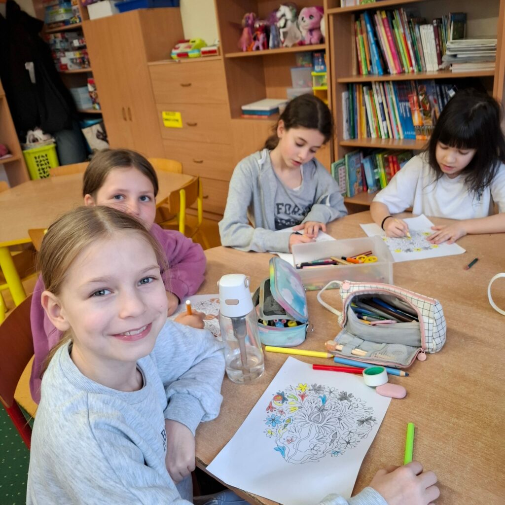 Na zdjęciu widać cztery dziewczynki siedzące przy stole w klasie i kolorujące rysunki z motywem kwiatowym. W tle znajdują się regały z książkami, zabawkami i grami, co sugeruje, że jest to przyjazne miejsce do nauki i zabawy. Na stole leżą przybory szkolne, takie jak piórniki, kredki i flamastry, co wskazuje na kreatywną pracę plastyczną. Jedna z dziewczynek na pierwszym planie uśmiecha się do aparatu, co nadaje zdjęciu radosny i przyjazny charakter.