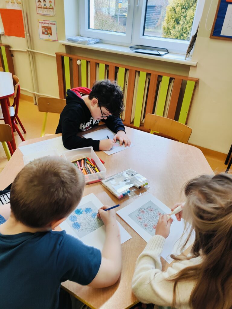 Na zdjęciu troje dzieci siedzi przy drewnianym stole w klasie i skupia się na kolorowaniu rysunków z motywem roślinnym. Każde z nich używa różnych kolorów, a na stole leżą kredki, mazaki i piórniki. W tle widać okno, przez które wpada naturalne światło, oraz elementy dekoracyjne klasy, takie jak plakaty edukacyjne. Atmosfera na zdjęciu jest spokojna i twórcza, co sugeruje, że dzieci cieszą się artystycznym zajęciem