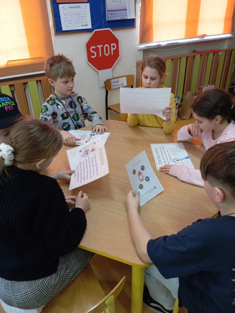 Na zdjęciu widać grupę pięciorga dzieci siedzących przy drewnianym stole w klasie. Każde z nich trzyma kartkę. Wszyscy czytają na temat dbania o zęby. W tle widać tablicę z kalendarzem, czerwony znak STOP oraz krzesełko z napisem „Usiądź. Wycisz się”. Klasa jest jasno oświetlona, a okna mają pomarańczowe rolety.