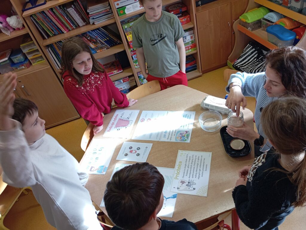 Grupa dzieci wraz z nauczycielką stoi wokół drewnianego stołu w klasie, obserwując eksperyment. Nauczycielka, ubrana w jasnoniebieski sweter, wkłada jajko do szklanego słoika z octem, a na stole leżą kartki z ilustracjami i tekstami na temat próchnicy, poprawnego szczotkowania zębów. W tle widać drewniane półki pełne książek, zabawek i przyborów szkolnych.