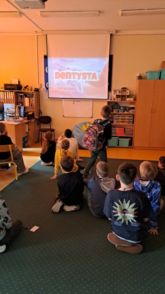 Grupa dzieci siedzi na dywanie w klasie i ogląda prezentację wyświetlaną na dużym ekranie, na którym widnieje napis "dentysta". W tle znajdują się półki z książkami, biurko z komputerem oraz szafki z materiałami szkolnymi.