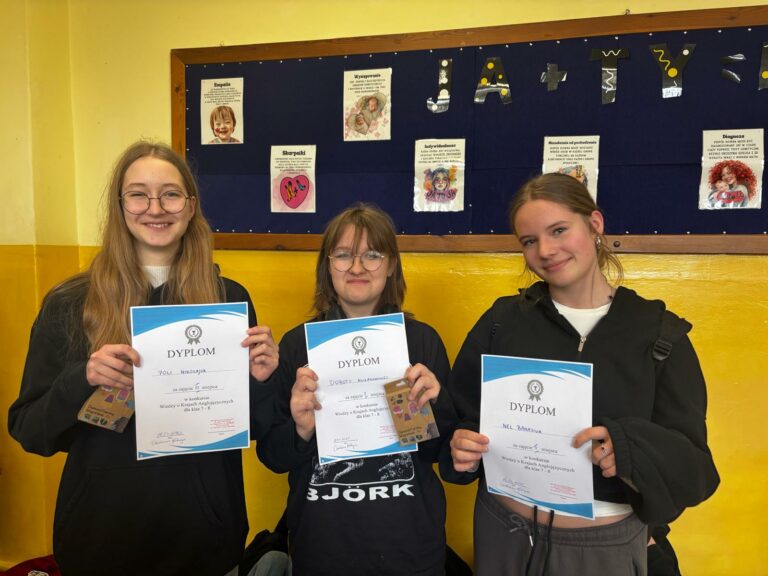 Na zdjęciu znajdują się trzy młode uśmiechnięte dziewczyny stojące obok siebie w pomieszczeniu szkolnym. Każda z nich trzyma w rękach dyplom za osiągnięcia w konkursie wiedzy o krajach anglojęzycznych dla klas 7. Dziewczyna po lewej ma długie, jasne włosy, nosi okulary i czarną bluzę. Trzyma dyplom za zajęcie III miejsca. Środkowa uczestniczka ma krótsze włosy, również nosi okulary i czarną bluzę z napisem „BJÖRK”. Również trzyma dyplom za II miejsce oraz pudełko. Dziewczyna po prawej ma włosy związane w niski kucyk, nosi czarną bluzę z kapturem i trzyma dyplom za zajęcie I miejsca.