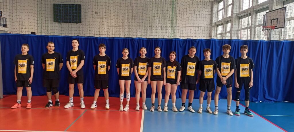 Zdjęcie przedstawia drużynę siatkówki, prawdopodobnie młodzieżową, ustawioną w równym szeregu na boisku. Zawodnicy ubrani są w jednakowe czarne koszulki z żółtym napisem „2004” oraz czarne spodenki. Niektórzy mają na nogach ochraniacze na kolana. Tło stanowi boisko do siatkówki z charakterystyczną siatką i niebieskimi kurtynami. Ułożenie zawodników sugeruje oficjalne zdjęcie drużynowe, być może przed lub po meczu. Atmosfera jest spokojna i skupiona, a wyrazy twarzy zawodników są neutralne. Zdjęcie jest ostre i dobrze naświetlone, co pozwala na wyraźne rozpoznanie szczegółów.