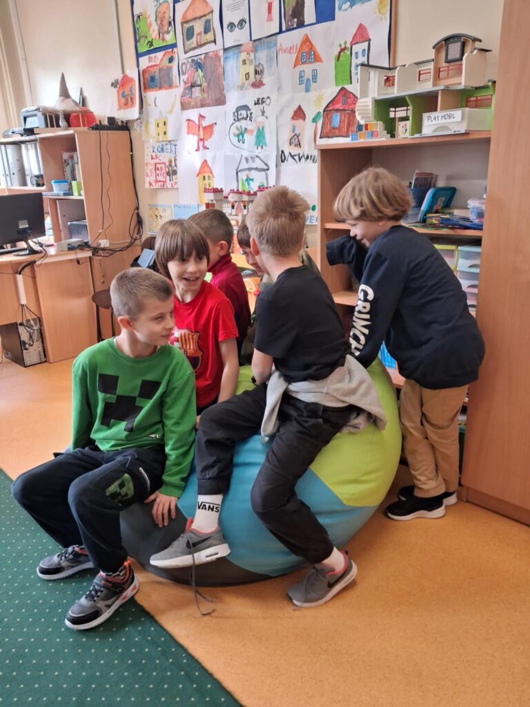 Na pierwszym planie widać czterech chłopców, siedzących na dużej kolorowej pufie. Za nimi stoi chłopiec, który opiera się o nią. Za nimi stoi regał z książkami i domkiem. Na tably wiszą kolorowe prace dzieci. Zdjecie zostało zrobione na świetlicy.