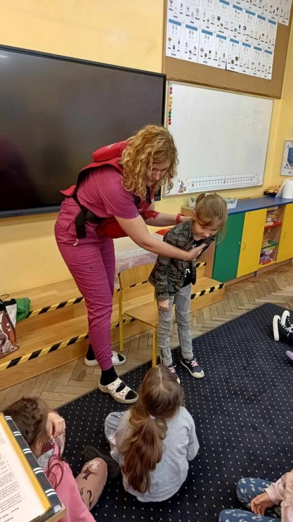 Na zdjęciu znajduje się kobieta w różowym stroju medycznym, która wykonuje na dziecku technikę pierwszej pomocy- manewr Heimlicha. Dziecko pochyla się do przodu, a kobieta kładzie jedną rękę na jego klatce piersiowej, a drugą na plecach, pokazując, jak udzielać pomocy w przypadku zadławienia. W tle widać inne dzieci siedzące na dywanie oraz klasę z tablicą, plakatami edukacyjnymi i kolorowymi szafkami.