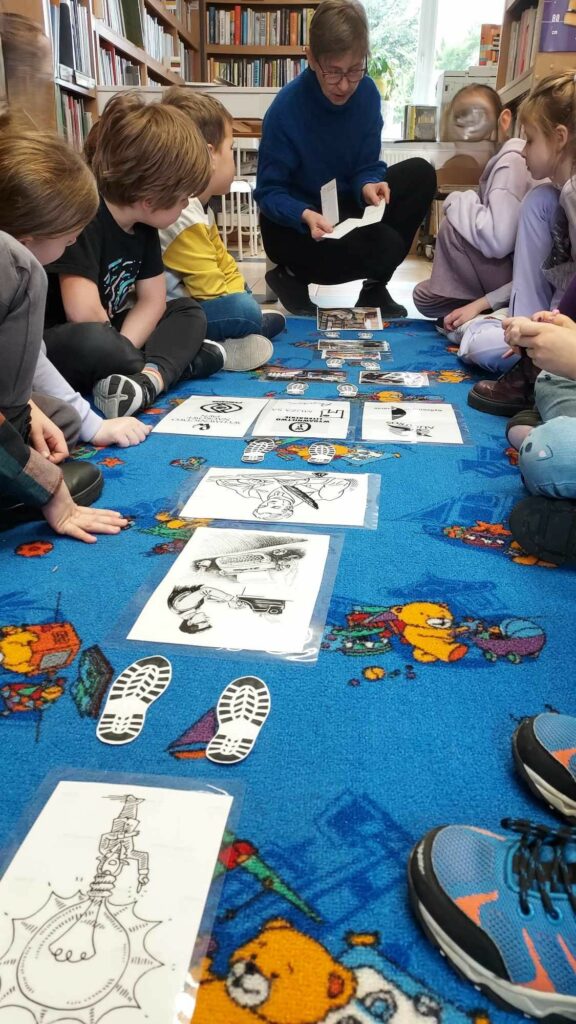 Zdięcie przedstawia grupę dzieci siedzących na niebieskim dywanie. Dzieci siedzą w dwóch grupach naprzeciw siebie pomiędzy nimi rozłożone są ilustracje oraz grafika śladów butów. Obok dzieci kuca kobieta trzymająca w ręku kartki. W tle widać regały z książkami.