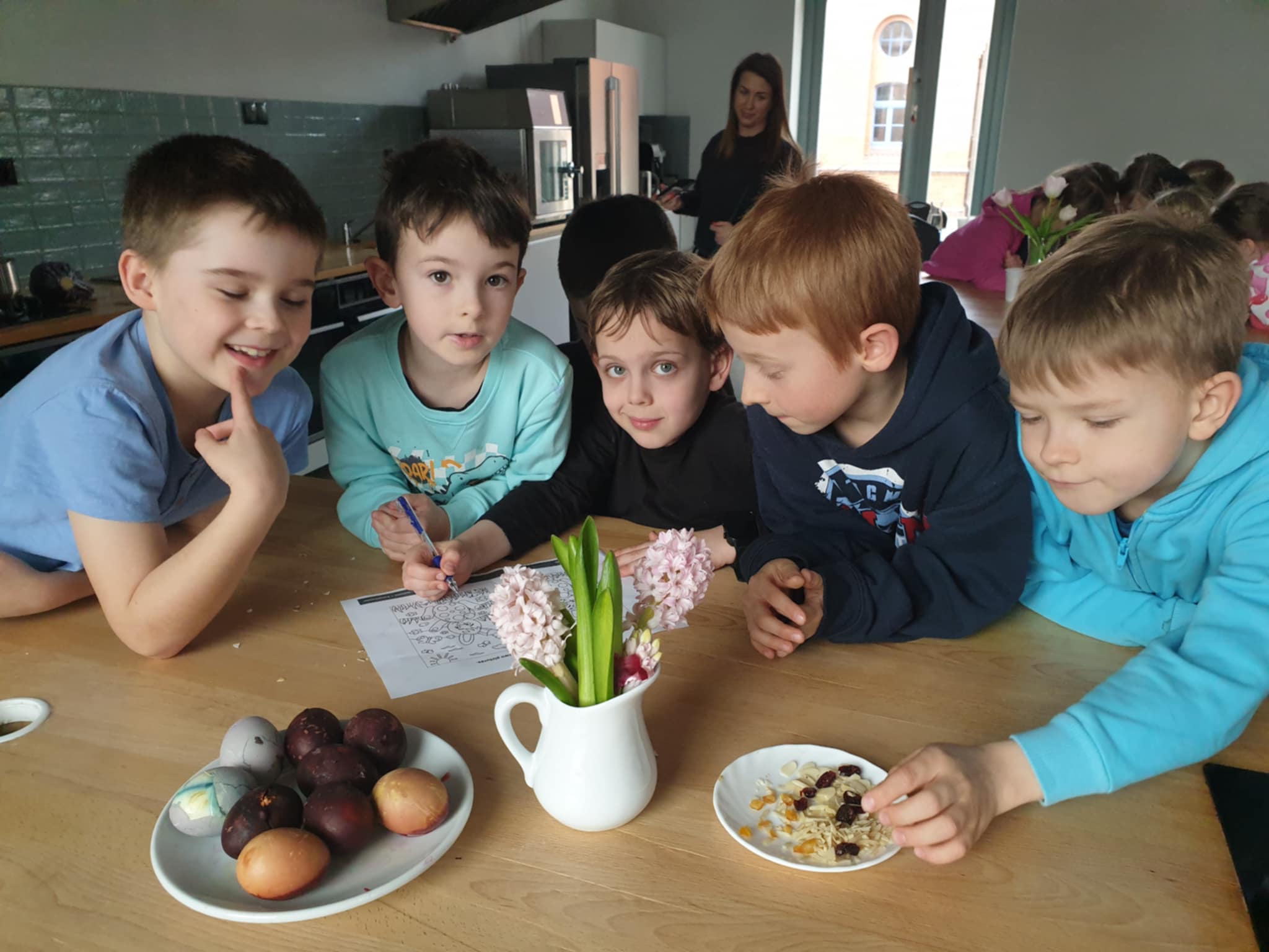 1. Na zdjęciu widać grupę pięciu chłopców siedzących przy drewnianym stole w jasnej kuchni. Dzieci wydają się być zaangażowane we wspólną aktywność – jedno z nich trzyma długopis i pisze na kartce, podczas gdy pozostali obserwują lub zajmują się jedzeniem. Na stole znajdują się talerze z barwionymi jajkami oraz płatkami z suszonymi owocami, a w tle można dostrzec inne dzieci oraz dorosłą kobietę.