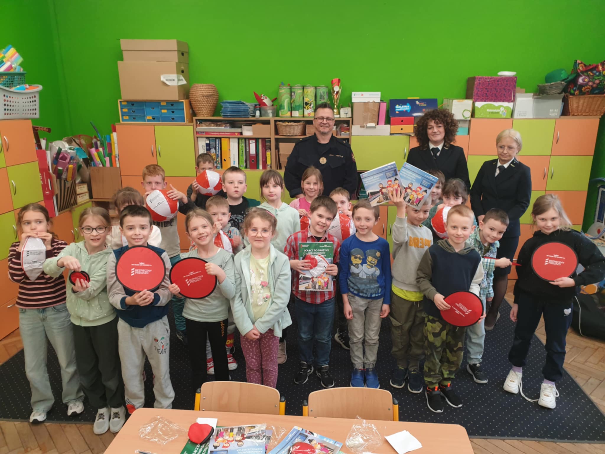 Na zdjęciu widzimy uczniów klasy 1a, którzy z uśmiechem pozują do wspólnego zdjęcia z przedstawicielami służb Kolei Państwowych. Dzieci trzymają w rękach różne materiały edukacyjne i gadżety, co wskazuje na przeprowadzone zajęcia o bezpieczeństwie. W tle widoczne są kolorowe szafki i przybory szkolne, co nadaje zdjęciu przyjazną i radosną atmosferę.